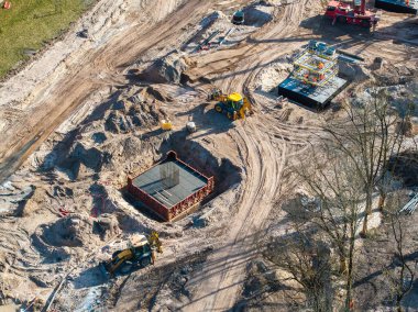 Riga, Letonya 'daki inşaat alanı, bölgede ağır makineler, toprak parçaları, beton yapılar, çelik iskeletler ve ağaçlar gösteriyor..