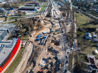 Riga, Letonya 'daki bir inşaat alanının hava görüntüsü, toprak çalışmalarını, ağır makineleri, mavi çadırları, kırmızı bir binayı ve çevresindeki yerleşim alanlarını gösteriyor..