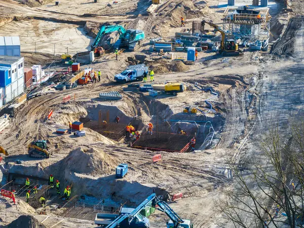 Yüksek görüş mesafesi olan işçiler, Riga şantiyesinde kazıcı ve vinç kullanıyorlar. Çelik yapı, eskrim ve şantiye ofisleri görülebilir durumda..