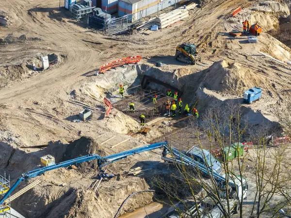Yüksek görüş mesafesi olan işçiler, Letonya 'nın Riga kentindeki bir inşaat sahasında altyapı inşa ediyorlar ve inşaat demirleri, mavi bir vinç ve güvenlik bariyerleri bulunuyor..