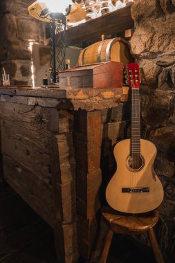Kırmızı başlıklı klasik bir gitar bir kereste çubuğunun yanındaki ahşap taburede duruyor. Taş bir duvar, fıçı ve sıcak bir lamba rahat bir alp ortamı yaratır..