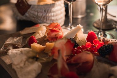 Peynirli ahşap tabaklar, kurutulmuş etler, ahududu, böğürtlen ve kırmızı frenk üzümü. Arka plandaki dokuma sepet ve şarap kadehleri güzel bir yemek olduğunu gösteriyor..
