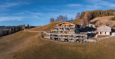 Güney Tyrol, İtalya 'da bir yamaçta çağdaş bir otel, arka planda cam pencereler, ahşap aksanlar, çimenli tarlalar ve kar kaplı dağlar var..