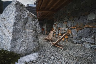 İki ahşap şezlong büyük bir kayanın yanında çakıl taşları üzerinde, Alp tarzında bir taş ve ahşap bina ve sakin bir ortamda zeminde kar..