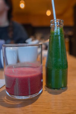 Bardakta kırmızı bir smoothie ve şişede yeşil bir meyve suyu ve tahta bir masada metal bir pipetle oturuyor. Sıcak ışıklandırma ve bulanık arkaplan samimi bir ortam önerir.