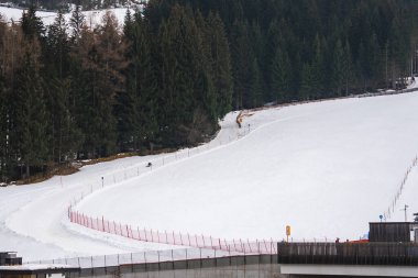 Yoğun yeşil ormanlarla çevrili, kırmızı güvenlik ağlarıyla çevrili bir kar yamacı. Ön planda modern bir yapı görülebilir..