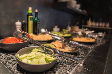 Dilimlenmiş salatalıklar, domatesler ve siyah taşlardaki siyah kaselerde sebzelerin yer aldığı bir büfe. Arka planda zeytinyağı ve sirke şişeleri var..