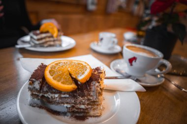 Beyaz bir tabakta turuncu bir dilim ile süslenmiş tiramisu yakın plan kahve fincanları, tatlılar ve kırmızı çiçekli saksı bitkisi..