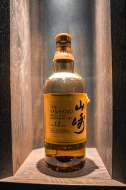 Bir şişe Yamazaki 12 yıllık Malt viskisi ahşap bir oyukta sergileniyor. Yumuşak ışıklandırmayla altın etiketi ve kehribar rengi sıvısı vurgulanıyor..