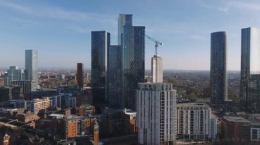 Manchester, İngiltere 'nin hava görüntüsü gökdelenleri, kırmızı tuğla binalar, yeşil alanlar, yollar ve açık mavi gökyüzü altındaki tren raylarını gösteriyor..