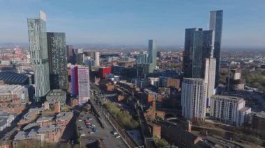 Manchester, İngiltere 'nin hava manzarası, gökdelenler, geleneksel binalar, tren rayları ve açık mavi gökyüzü altında hareket eden bir tren..