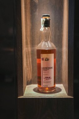 18 yıllık bir AnCnoc viski şişesi yumuşak ışıklandırmayla ahşap bir oyukta oturur, kehribar rengi ve minimalist etiket tasarımı sergilenir..