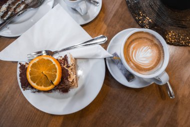 Beyaz bir tabakta turuncu garnitürlü bir tiramisu dilimi, latte sanatıyla eşleştirilmiş, lüks bir ortamda ahşap bir masaya kurulmuş..