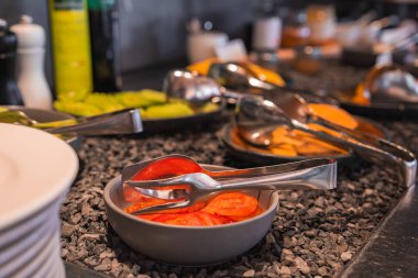 Lüks bir otelde, siyah taş bir yüzey üzerinde meyveler ve sebzelerle çevrili, maşalı bir kase dilimlenmiş domatesin yer aldığı bir büfe..