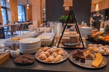 Modern bir yemek alanında hamur işleri, meyve ve yoğurt ile kahvaltı. Tahta paneller, büyük pencereler ve dışarıda karla kaplı ağaçlar var..