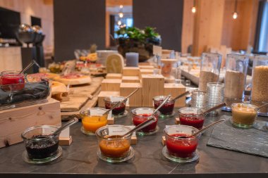 Ahşap tahtaların üzerinde reçel, mısır gevreği, ekmek ve hamur işleri olan şık bir kahvaltı büfesi, yumuşak ışıklandırmalı sıcak ahşap bir iç mekâna kurulmuş..