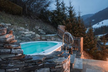 İtalyan Alpleri 'ndeki alacakaranlık gökyüzünün altında yeşil ağaçlar ve kar kaplı yamaçlarla çevrili kırsal taş işçiliğiyle aydınlatılmış bir açık hava jakuzisi..