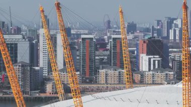 O2 Arena 'nın havadan görünüşü, sarı direkler, modern yüksek binalar, vinçler ve Thames Nehri. Yavaş yavaş Londra 'nın gelişen ufuk çizgisini gözler önüne seriyor.