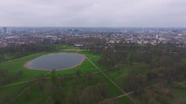 Oval bir göl, yürüyüş yolları ve yeşillik içeren bir Londra parkının havadan görünüşü. Kademeli zoom modern ve tarihi binalarla şehir manzarasını gözler önüne seriyor.