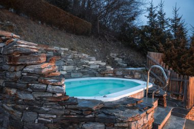 Bir yamaçta kırsal bir taşın içine kurulmuş açık bir jakuzi. Etrafı yemyeşil ağaçlar ve eğimli arazilerle çevrili. İtalyan Alpleri 'nde alacakaranlıkta ele geçirilmiş..