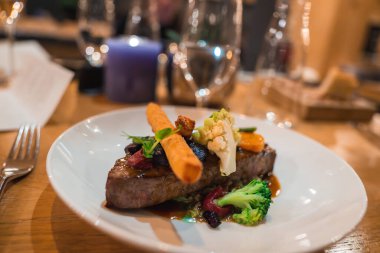 Kızarmış havuç, karnabahar ve brokoli ile kaplanmış biftek, beyaz bir tabakta. Zarif cam eşyalar ve yanan mor bir mum atmosferi güçlendirir..