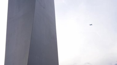 Parlak gökyüzüne karşı uzun, parlak, modern bir dikilitaş. Kamera hafifçe yana yatar ya da döner, yapının yüksekliğini ve tasarımını gösterir.