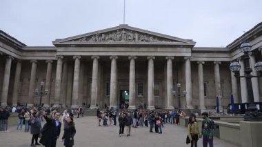 Ziyaretçiler, bulutlu gökyüzünün altındaki kalabalık avluda yürürken, sohbet ederken ve fotoğraf çekerken, İngiliz Müzesi 'nin sütunları ve heykelleriyle neoklasik cephesi.