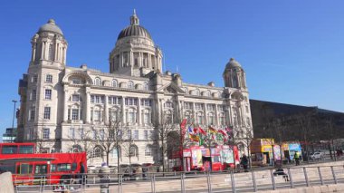 Liverpool Limanı, Liverpool 'da açık mavi bir gökyüzünün altında duruyor. Kırmızı otobüsler, yemek tezgahları, yayalar ve hafif trafik olay yerine hareket getiriyor..
