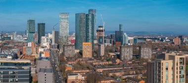 Manchester, İngiltere 'nin hava görüntüsü, modern gökdelenler, inşaat vinçleri, yerleşim alanları, sokaklar, yeşillikler ve açık bir gökyüzü altında yuvarlanan tepeler.