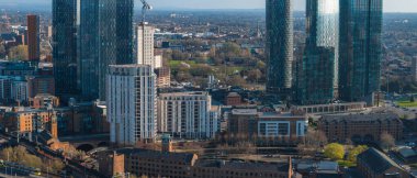 Manchester, İngiltere 'nin hava görüntüsü, modern gökdelenleri, tarihi tuğla binaları, bir demiryolu hattı, yeşil alanlar ve güneşli bir günde uzak tepeleri gösteriyor..