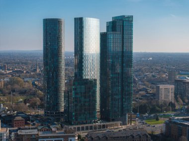 Manchester, İngiltere 'nin hava manzarası, daha küçük binalar, yeşillik ve açık mavi gökyüzü ile çevrili üç yansıtıcı cam gökdelenler içerir..