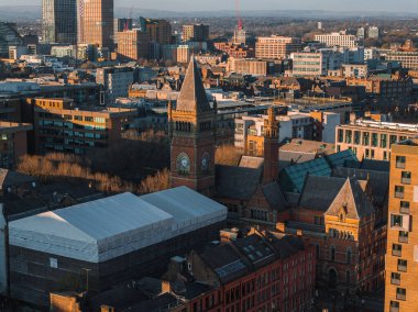 Manchester, İngiltere 'nin hava görüntüsü, tarihi kırmızı tuğlalı saat kulesini, modern gökdelenleri, çeşitli mimariyi, açık gökyüzünü ve uzak yeşillikleri gösteriyor..