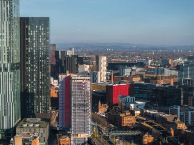 Manchester, İngiltere 'nin hava manzarası, gökdelenler, orta yükselen binalar, demiryolu rayları ve açık mavi gökyüzü altındaki yuvarlanan tepeler.