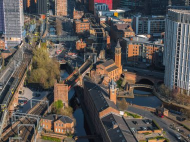 Güneşli bir günde Manchester, İngiltere 'nin hava görüntüleri kırmızı tuğlalı binaları, kanalları, modern gökdelenleri ve görünür trenleri olan bir demiryolu hattını gösteriyor..