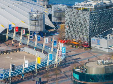 O2 Arena 'nın beyaz kubbesi ve direkleri modern binalarla çevrili, arkasında pankartlar olan bir yürüyüş yolu ve arkasında Thames Nehri var..