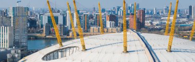 O2 Arena 'nın beyaz kubbesi ve sarı direkleri modern yüksek binaların ortasında, Thames Nehri ve Canary Rıhtımı arka planda görünür..