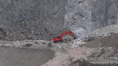 Alplerdeki bir maden alanının havadan görüntüsü, turuncu bir kazıcının kaya kırdığını, enkaz yığınları yarattığını ve dik kayalıkların arasında işlediğini gösteriyor..