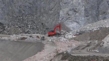 Alplerdeki bir maden sahası havadan görünüyor. Kırmızı bir kazıcı moloz yığınlarının yakınındaki enkazı hareket ettiriyor. Toprak yollar ve engebeli arazilerle çevrili..