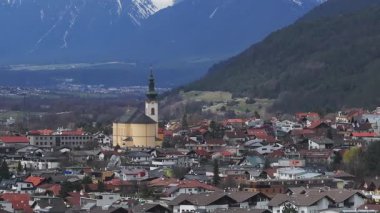 Alplerdeki bir kasabanın havadan görünüşü sarı bir kilise ve yüksek bir kule, kırmızı çatılı binalar ve karla kaplı dağlar pürüzsüz bir şekilde hareket ediyor..