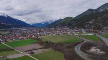 Alplerdeki bir vadinin hava manzarasında küçük bir kasaba, kırmızı çatılı binalar, yemyeşil tarlalar, dolambaçlı bir nehir ve dağ zirveleri olan ormanlık tepeler var..