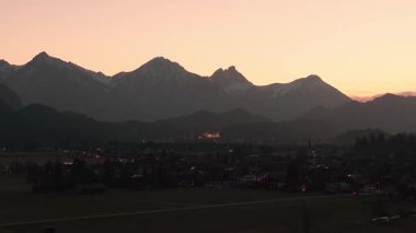 Neuschwanstein Kalesi 'nin hava manzarası gün batımında aydınlandı. Bavyera Alpleri ve çerçevesinde bir kilise vardı. Gökyüzü alacakaranlığa dönüşür.