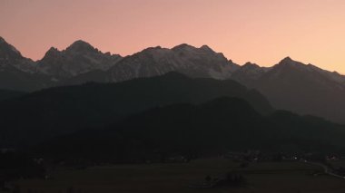 Almanya, Bavyera 'daki Neuschwanstein Kalesi' nin günbatımında havadan görüntüsü. Dağ zemin arkaplanı, virajlı yol ve köy ışıklarıyla aydınlatılmış bir kale..