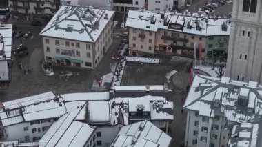 Kışın Cortina d 'Ampezzo' nun hava görüntüsü, karla kaplı çatılar, beyaz bir çan kulesi, yeşil bir çatı kilisesi ve yayaların olduğu bir kasaba meydanı gösteriyor..