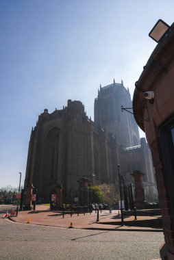 Liverpool Katedrali açık mavi gökyüzünün altında Gotik Uyanış tasarımı ile dimdik ayakta duruyor. Ön planda asfalt, kalın ve park halindeki arabalar görünüyor..