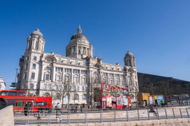 İngiltere 'deki Liverpool Limanı binasında Edwardian Barok mimarisi, kırmızı çift katlı otobüsler, bayraklı turist kulübeleri ve açık mavi bir gökyüzü yer almaktadır..