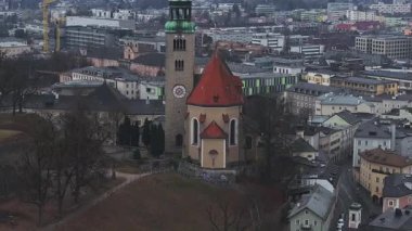 Bulutlu bir günde, şehir binaları, yollar ve köprülerle çevrili, kırmızı kiremitli çatısı ve yeşil kubbesi olan Salzburg 'daki tarihi bir kilisenin hava manzarası..