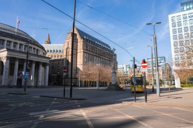 Manchester sokak manzarasında dairesel Merkez Kütüphanesi, modern binalar ve açık mavi gökyüzünün altında sarı bir Metrolink tramvay var..