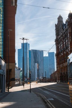 Manchester sokak manzarasında kırmızı tuğlalı tarihi mimari modern Deansgate Meydanı gökdelenleri, tramvay rayları ve açık mavi gökyüzü var..
