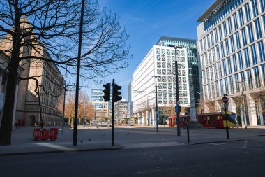 Solunda taş bina olan bir Manchester caddesi manzarası, sağında cam yapılar, kırmızı bir otobüs, çıplak ağaçlar, bir trafik ışığı ve açık mavi bir gökyüzü..
