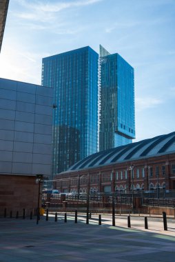 Manchester manzarasında Deansgate Meydanı kuleleri, tarihi bir kırmızı tuğla bina, kaldırımlı bir alan ve açık mavi bir gökyüzü var..